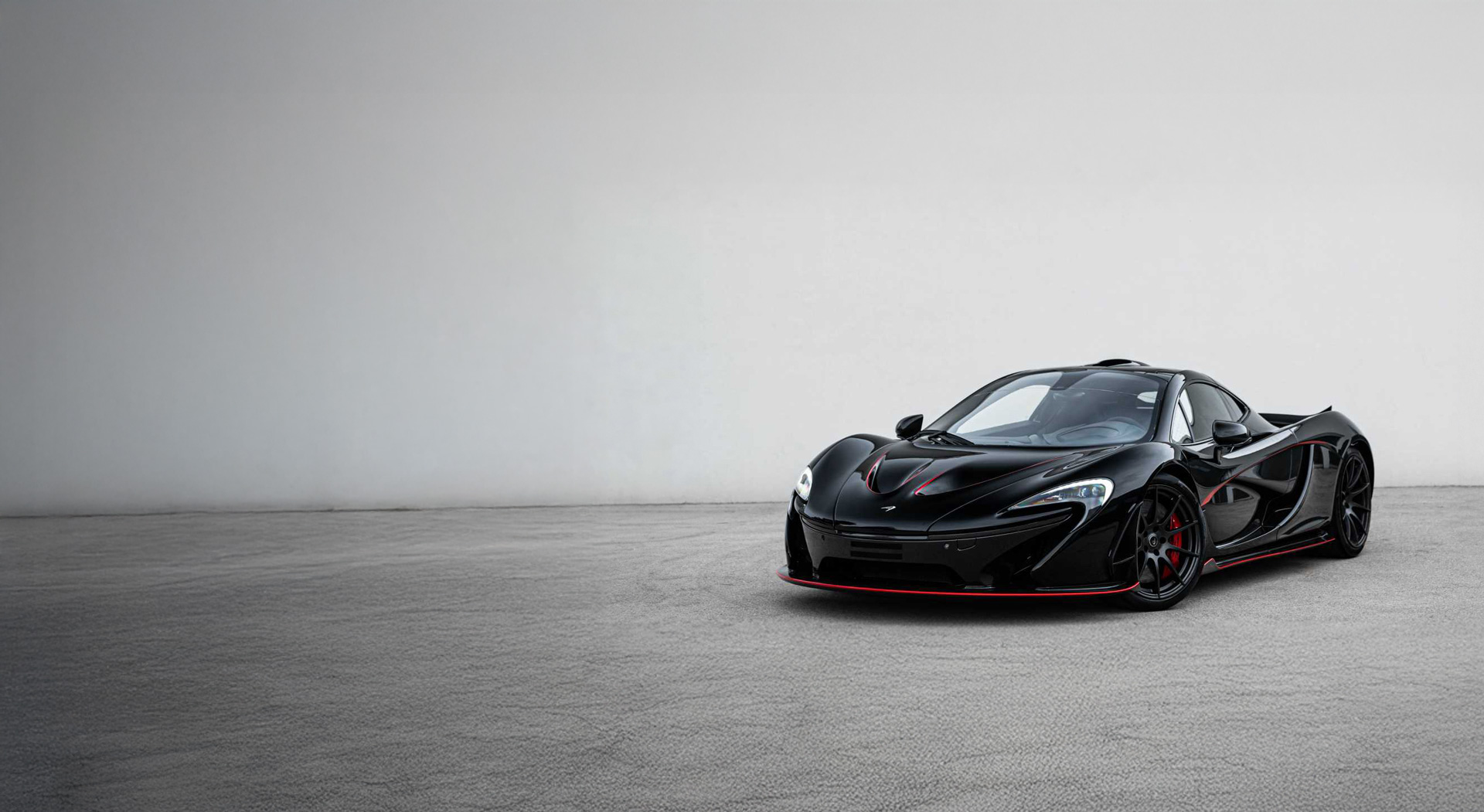 Black McLaren P1 with red detailing, parked on tarmac in front of white wall.